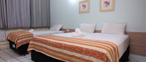 A hotel room with two beds, each with a striped bedspread and pillows. There are two framed pictures on the wall, a window with curtains, and a tiled floor.