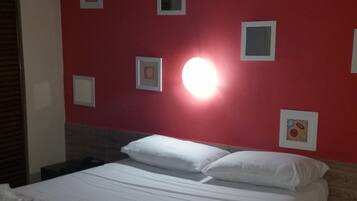 Uma cama bem arrumada com lençóis brancos e um edredom com padrão floral, uma parede vermelha com quadros emoldurados, e uma fonte de luz clara acima da cama.