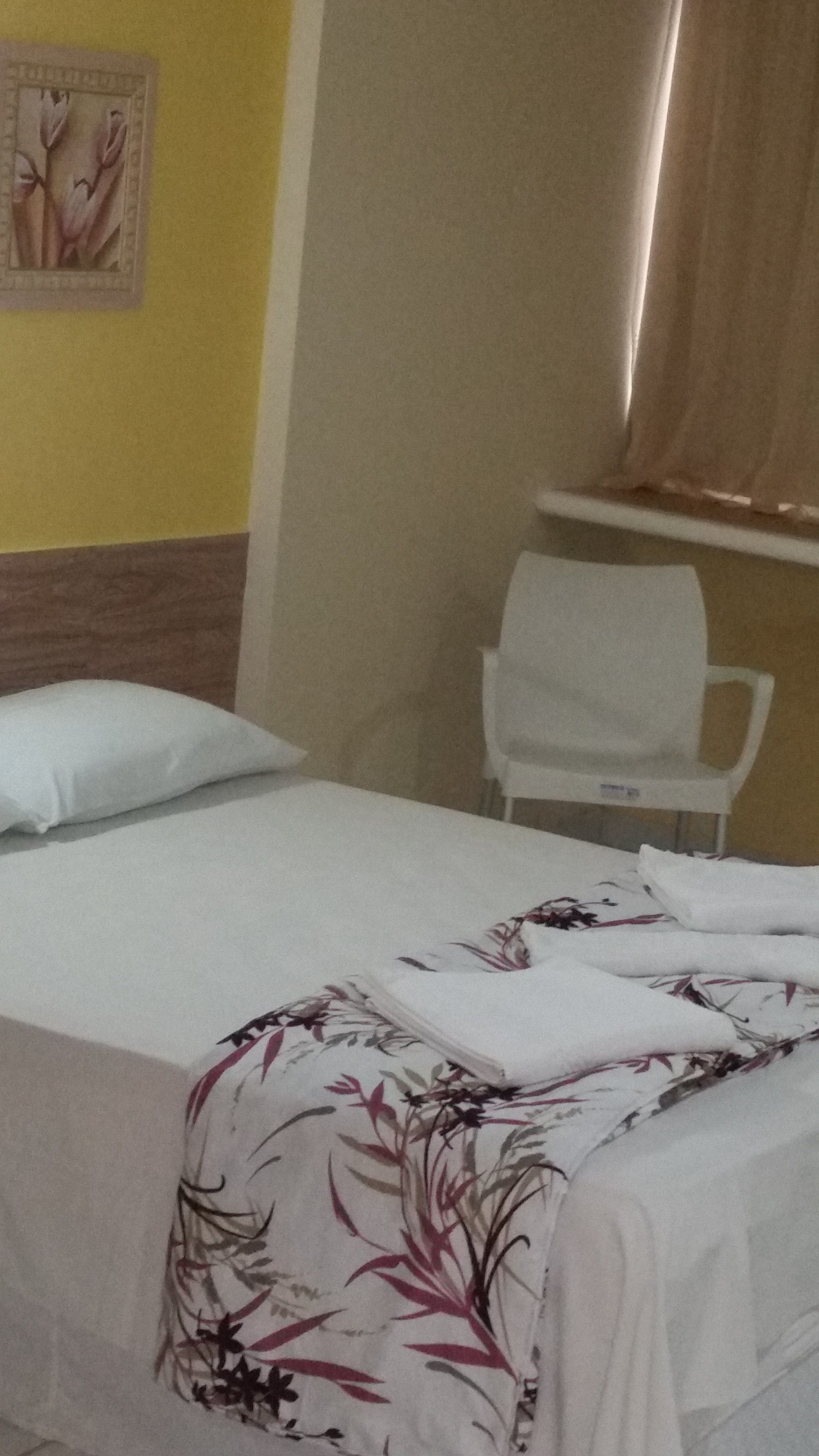 A single bed with a floral bedspread, a white pillow, and a plain wall with a framed picture.