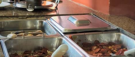 Una barra libre con distintos alimentos en fuentes de metal, incluyendo carnes y arroz.