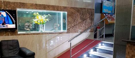 A lobby with a marble wall, a black leather sofa, a glass table with red balls, a red carpeted staircase, and a digital screen.