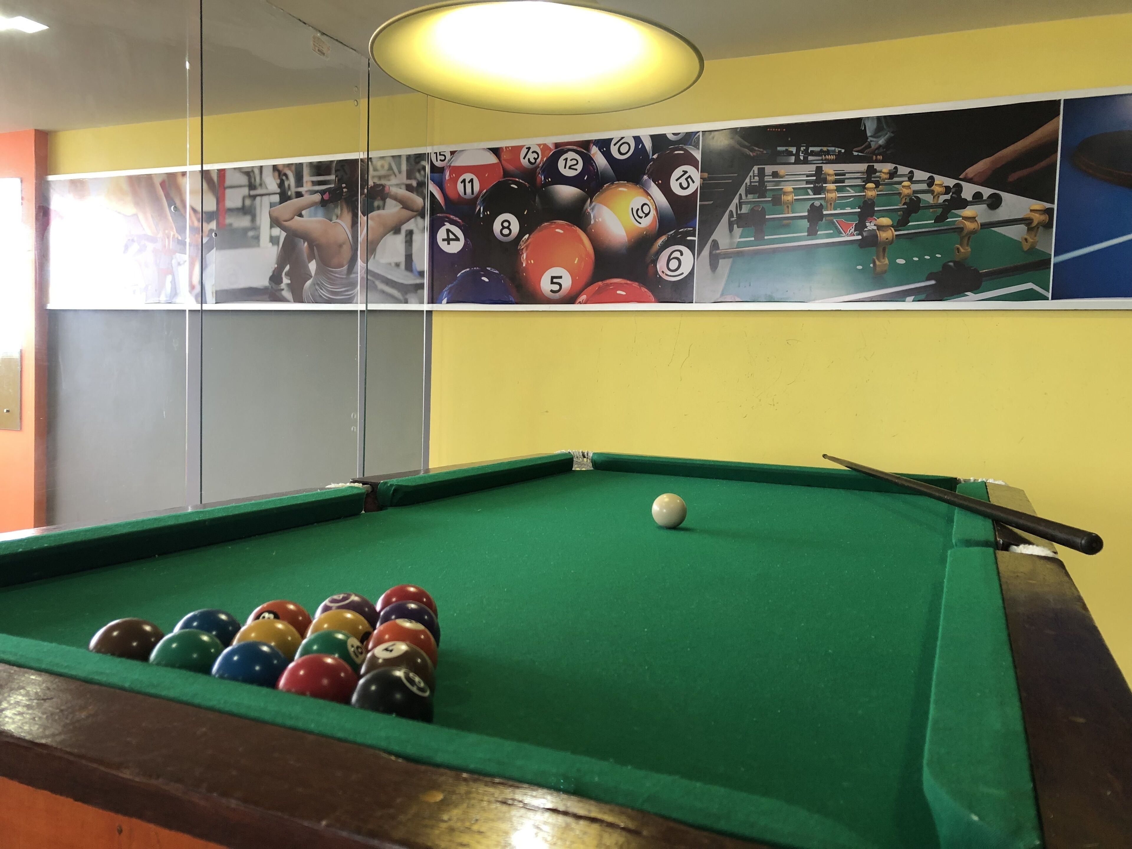 Une table de billard avec des billes et une queue, une cloison en verre et un mur orné d’images de sport.