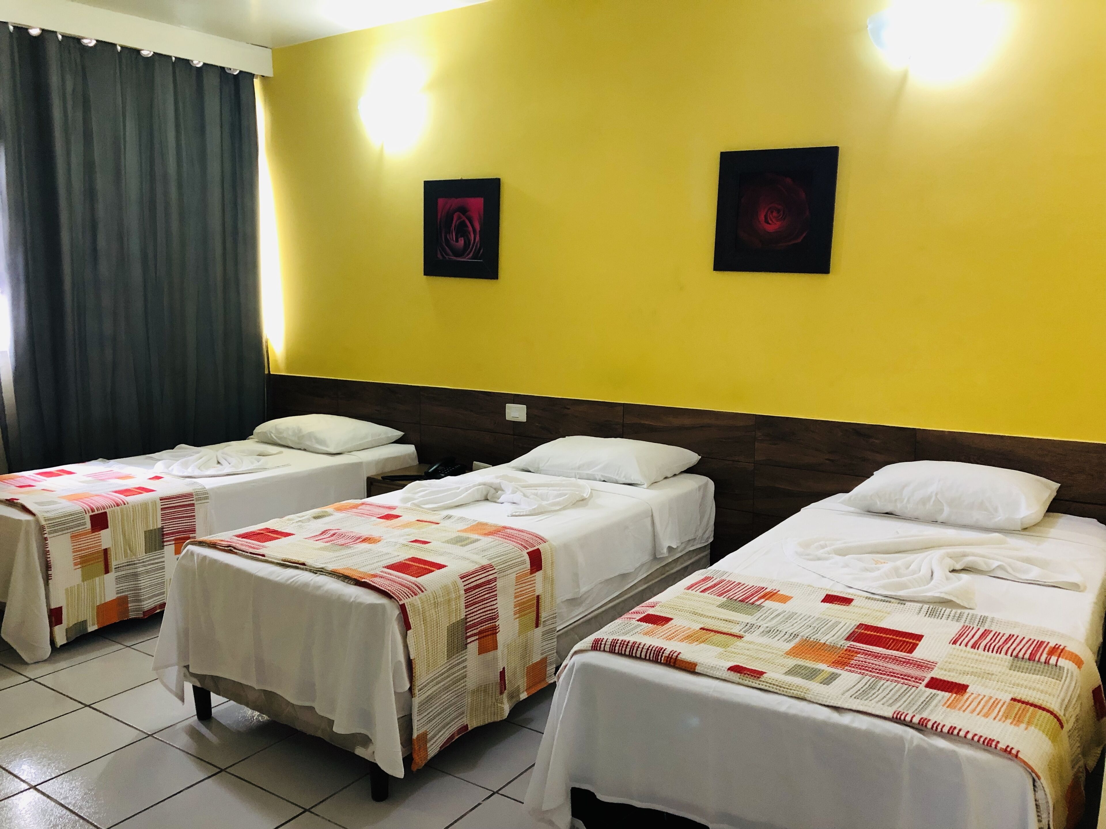 Quarto de hotel com três camas de solteiro, cada uma com colcha branca e edredom colorido. O quarto tem paredes amarelas, cabeceira de madeira escura e duas molduras com quadros na parede.