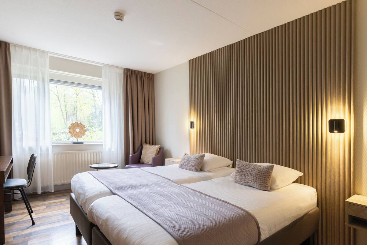 A hotel room with two beds, a desk, a chair, a window with curtains, and a wall with vertical blinds.