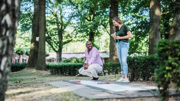 A person playing shuffleboard in an outdoor setting with trees and a paved area.