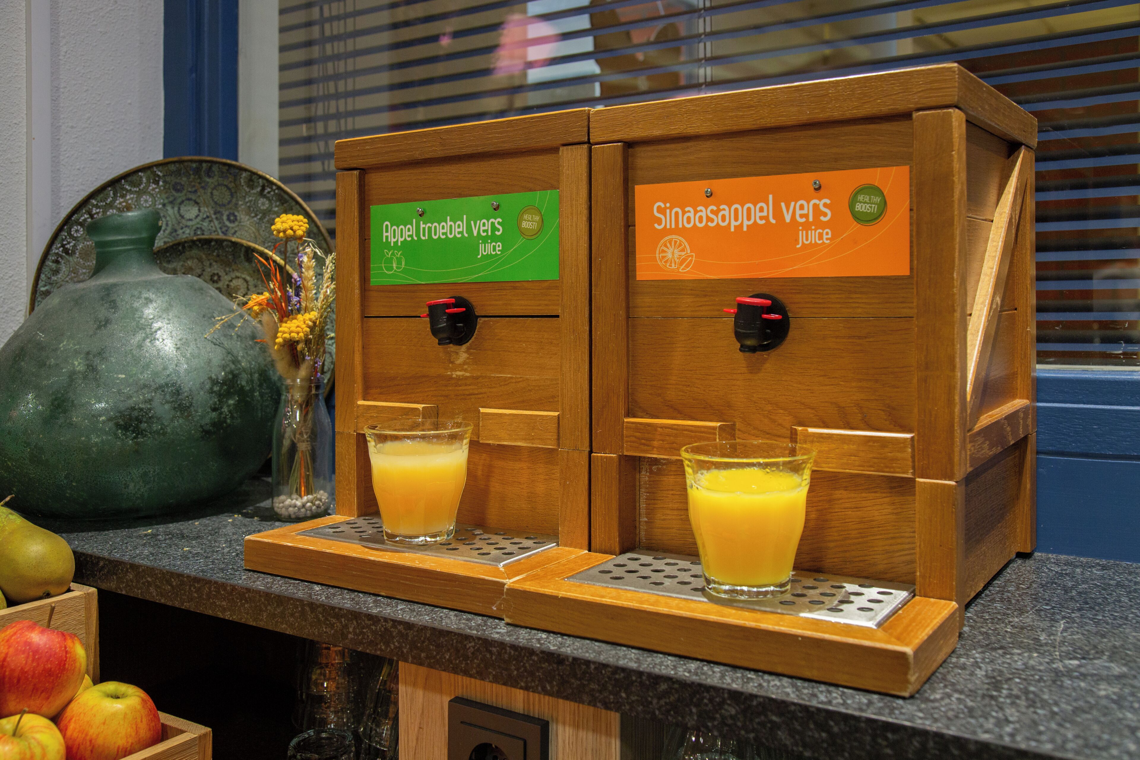 A juice dispenser with two types of juice, a green label for 'Appel troebel vers juice' and an orange label for 'Sinaasappel vers juice', and a decorative vase with yellow flowers on the counter.