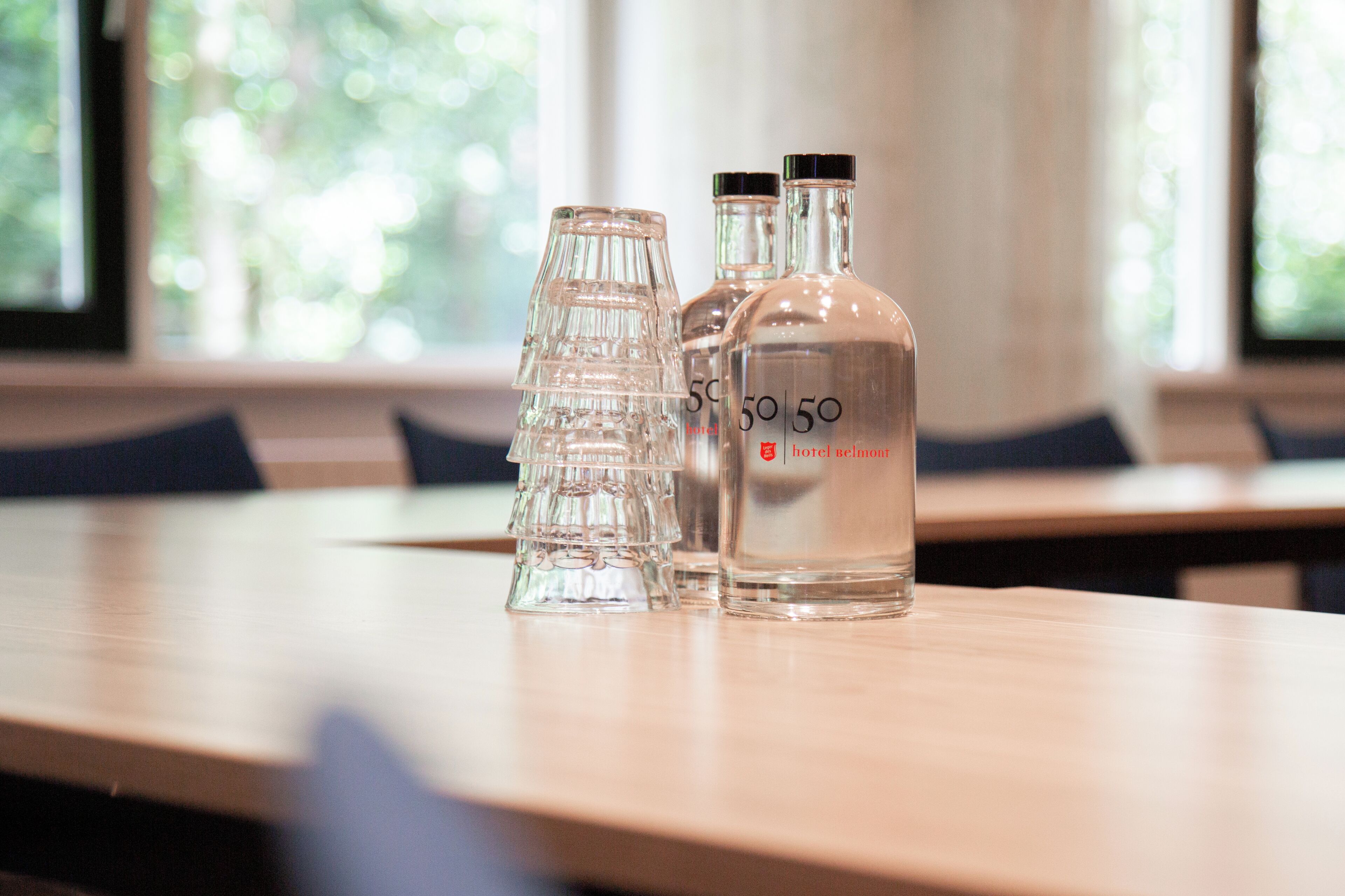 Una mesa con tres botellas de vidrio transparente y una jarra de vidrio, probablemente con agua, en una habitación con vista a árboles a través de una ventana.