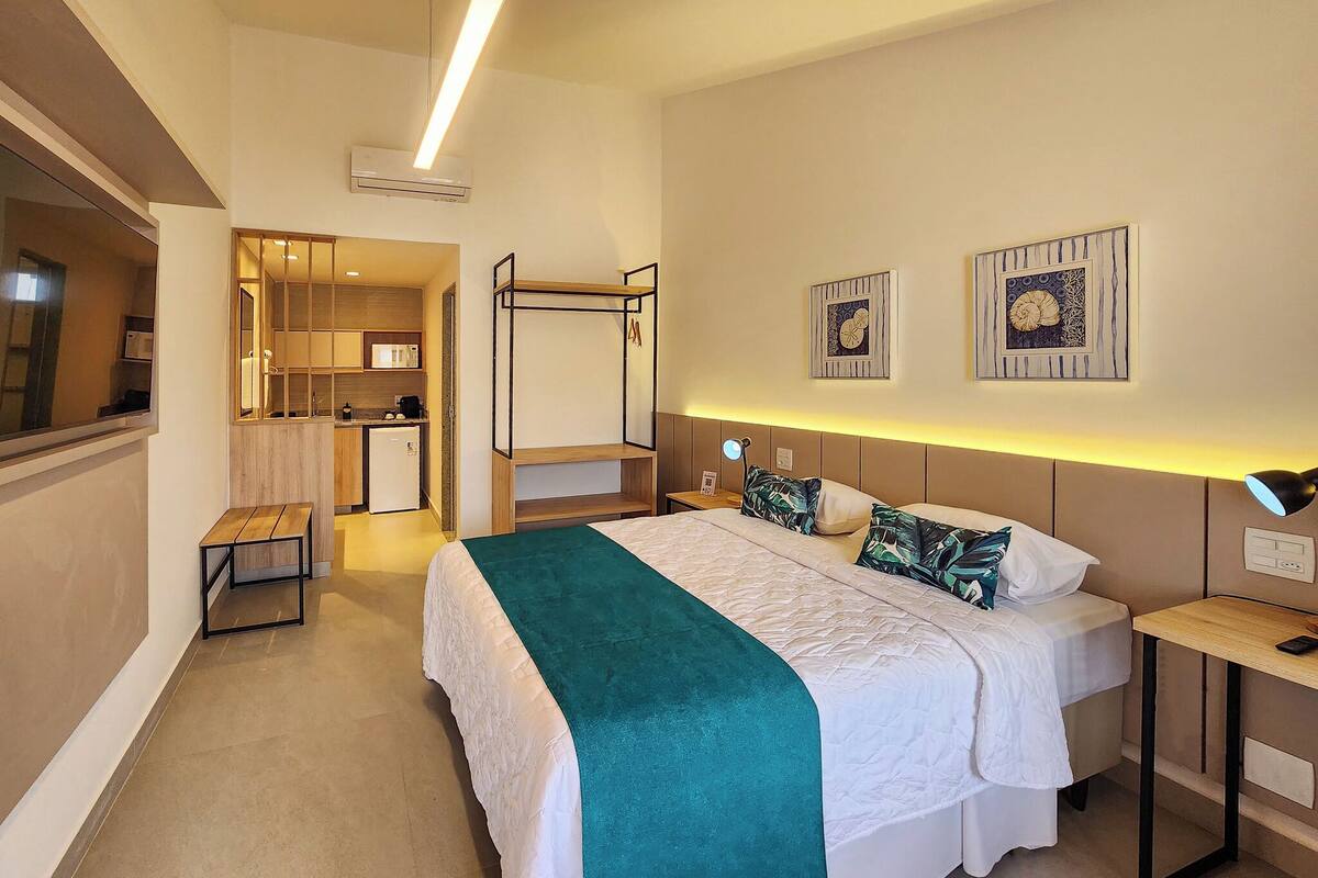 A hotel room with a bed, bedside table, and a small desk. There is a kitchenette area with a microwave and a refrigerator.