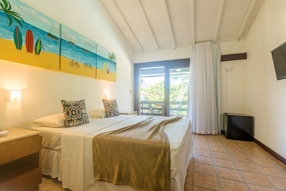 A hotel room with a bed, a painting of a beach scene, a window with curtains, and a television on the wall.