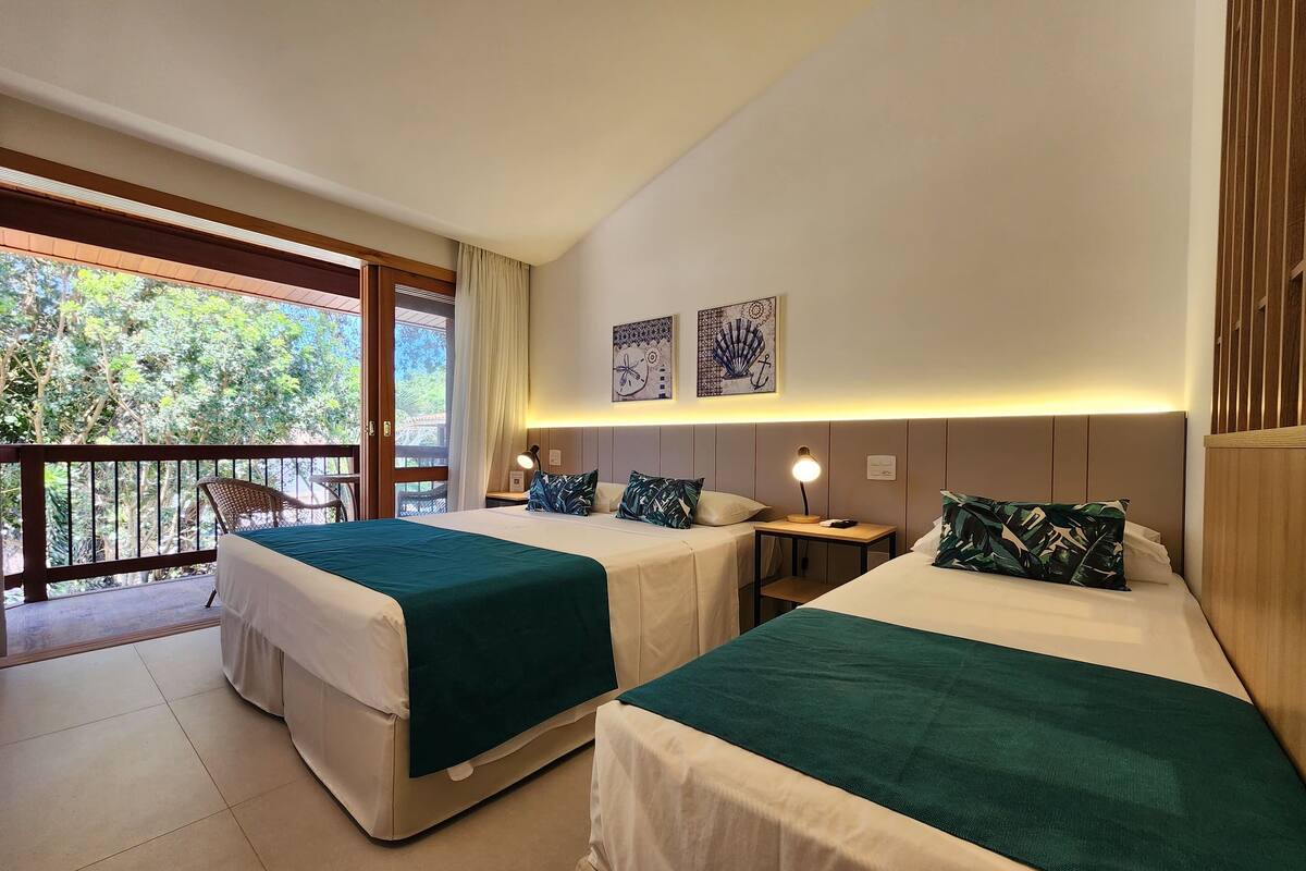 A hotel room with two beds, a balcony with a view of trees, and a small table with a lamp.
