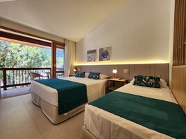 Quarto de hotel com duas camas, varanda com vista para árvores e uma pequena mesa com abajur.