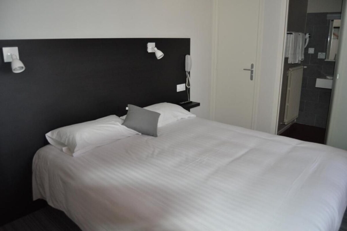 Una cama bien tendida con sábanas blancas y una sola almohada, una mesita de noche con lámpara, y una puerta que da a un baño con espejo y perchero.