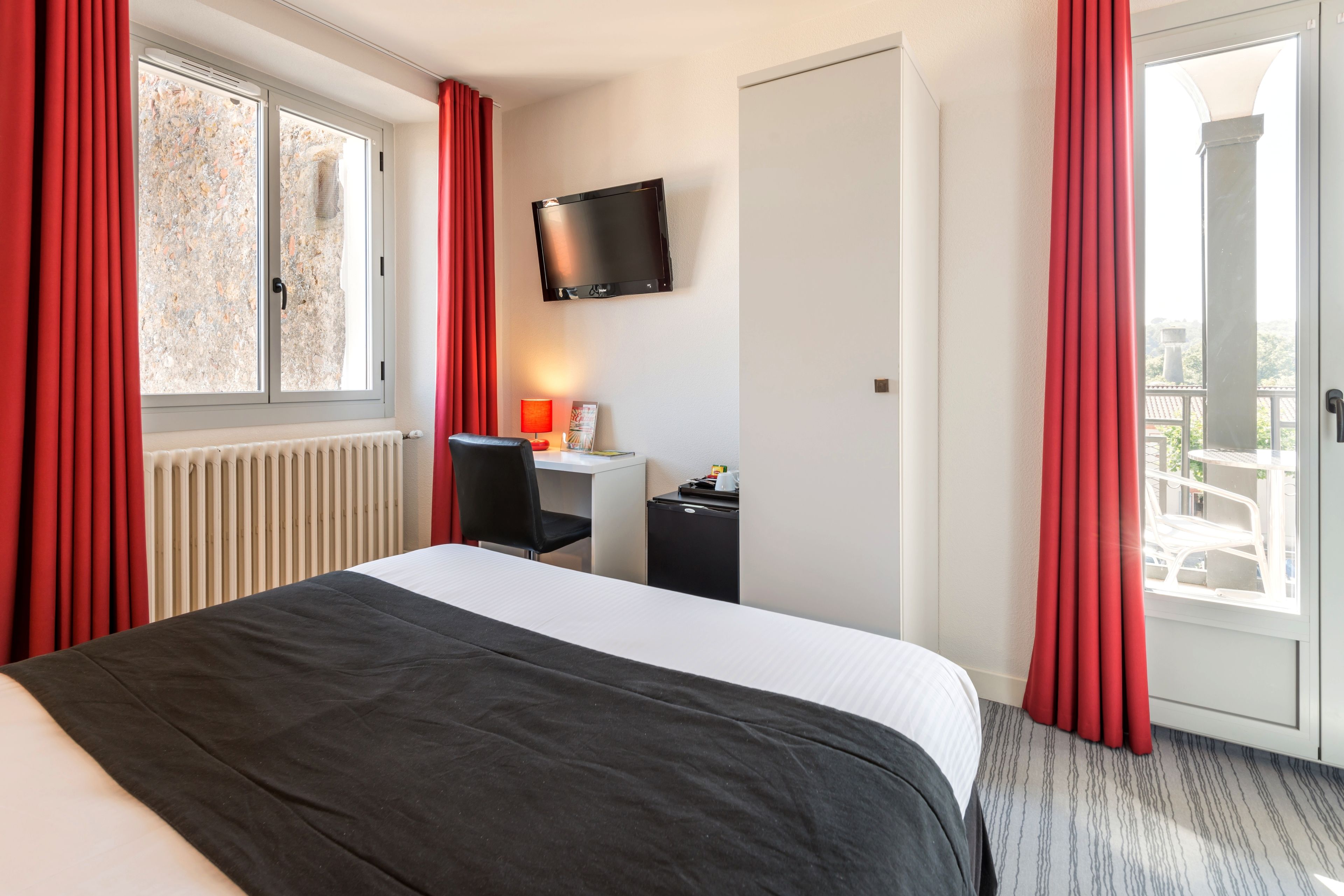 Una camera d'albergo con un letto, una scrivania, una sedia, una televisione e una finestra con tende rosse.
