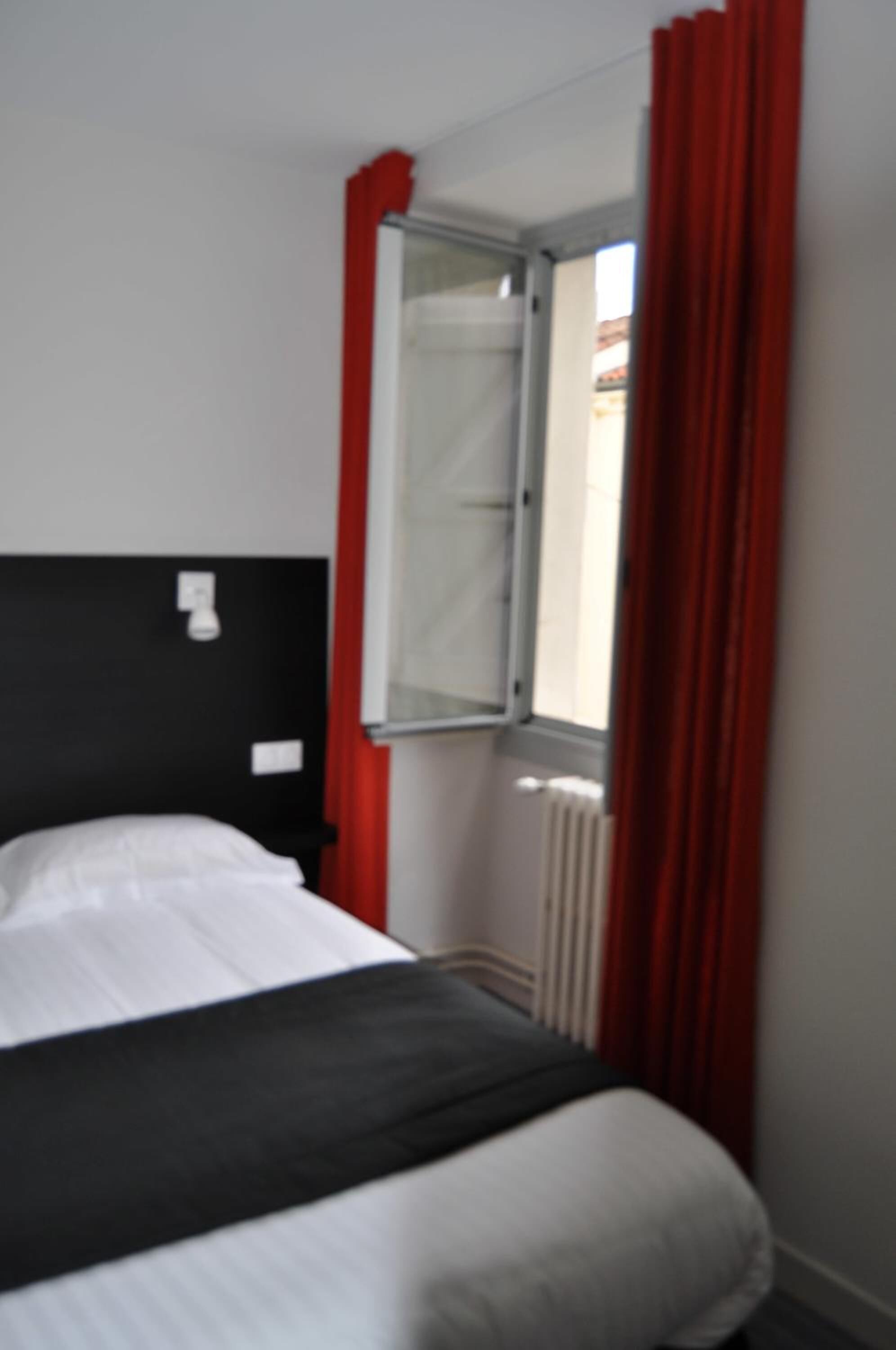 A hotel room with a bed, a window with red curtains, and a white wall.