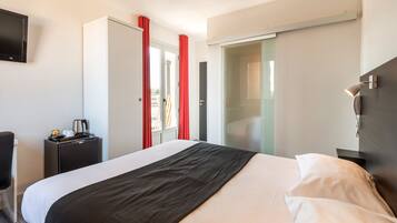 A hotel room with a bed, a desk with a coffee maker, a TV, and a sliding door with red curtains.