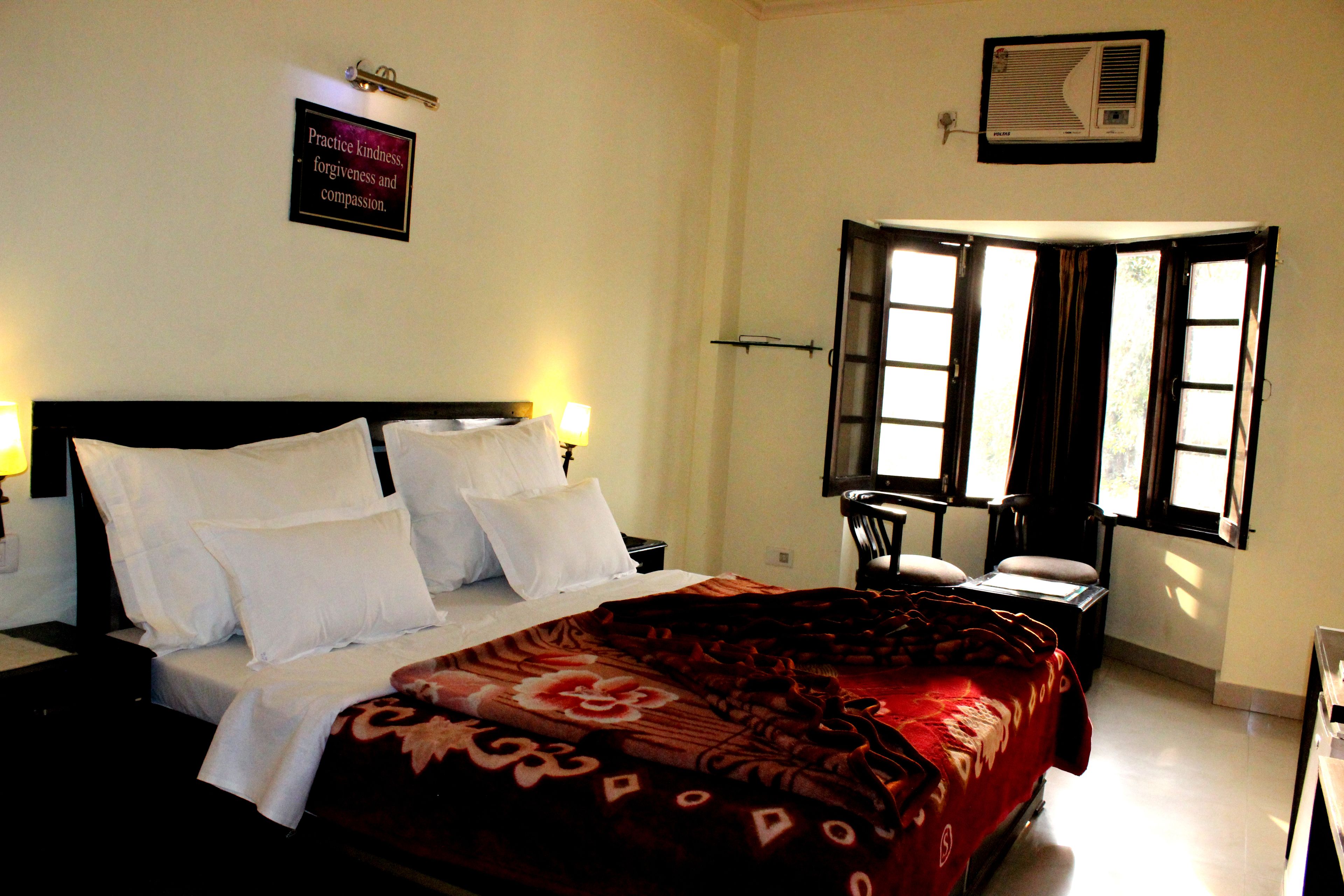 A hotel room with a bed, bedside tables, a desk, and a chair. There is a window with curtains and a wall-mounted air conditioner.