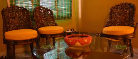 A room with two ornate wooden chairs with cushioned seats, a glass coffee table with a decorative bowl, and a window with blinds.