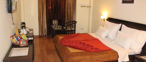 A hotel room with a wooden bed, a red blanket, a desk with snacks, a chair, and a curtain.