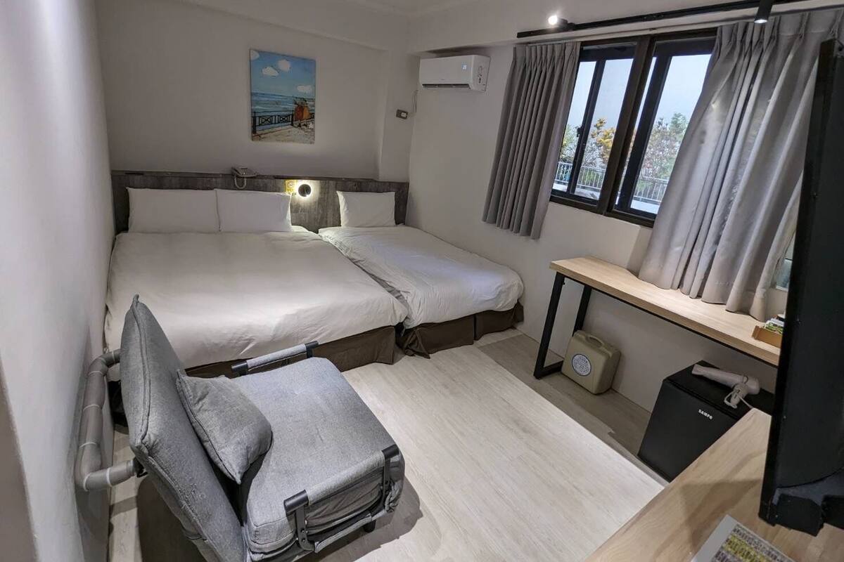 Una habitación de hotel pequeña con cama, escritorio, silla, ventana con cortinas, unidad de aire acondicionado y un cuadro en la pared.