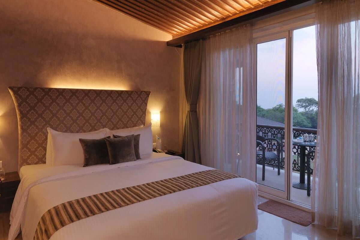 A hotel room with a large bed, a nightstand, a wall-mounted lamp, and a balcony with outdoor furniture.
