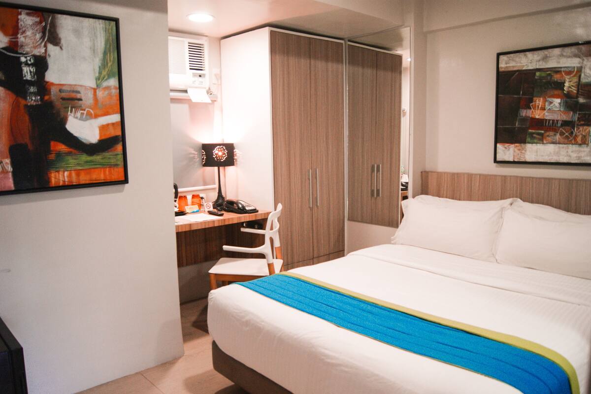 A hotel room with a bed, a desk, a chair, a wardrobe, and two paintings on the walls.
