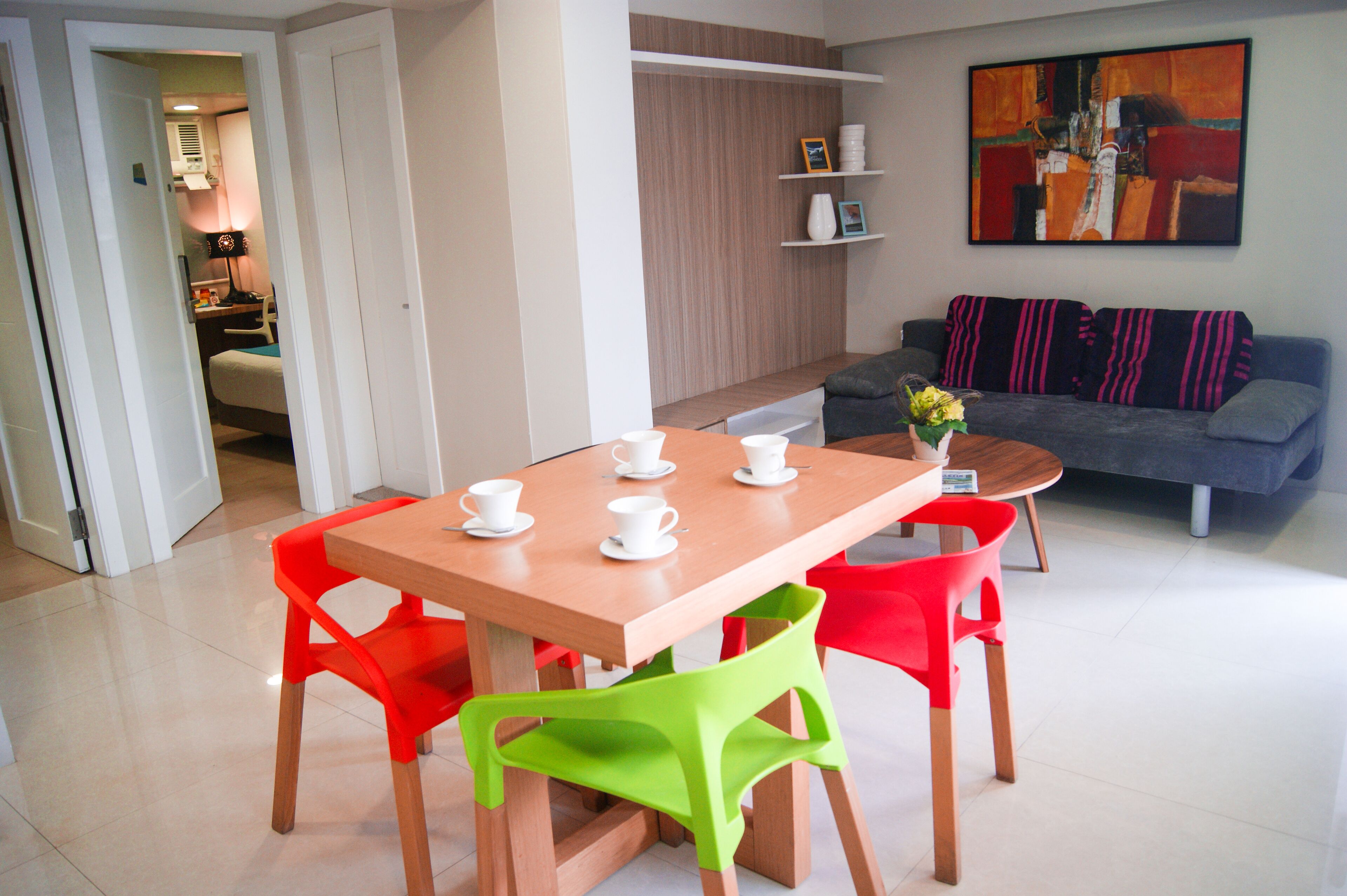 A modern living room with a wooden dining table, red and green chairs, a grey sofa, and a painting on the wall.
