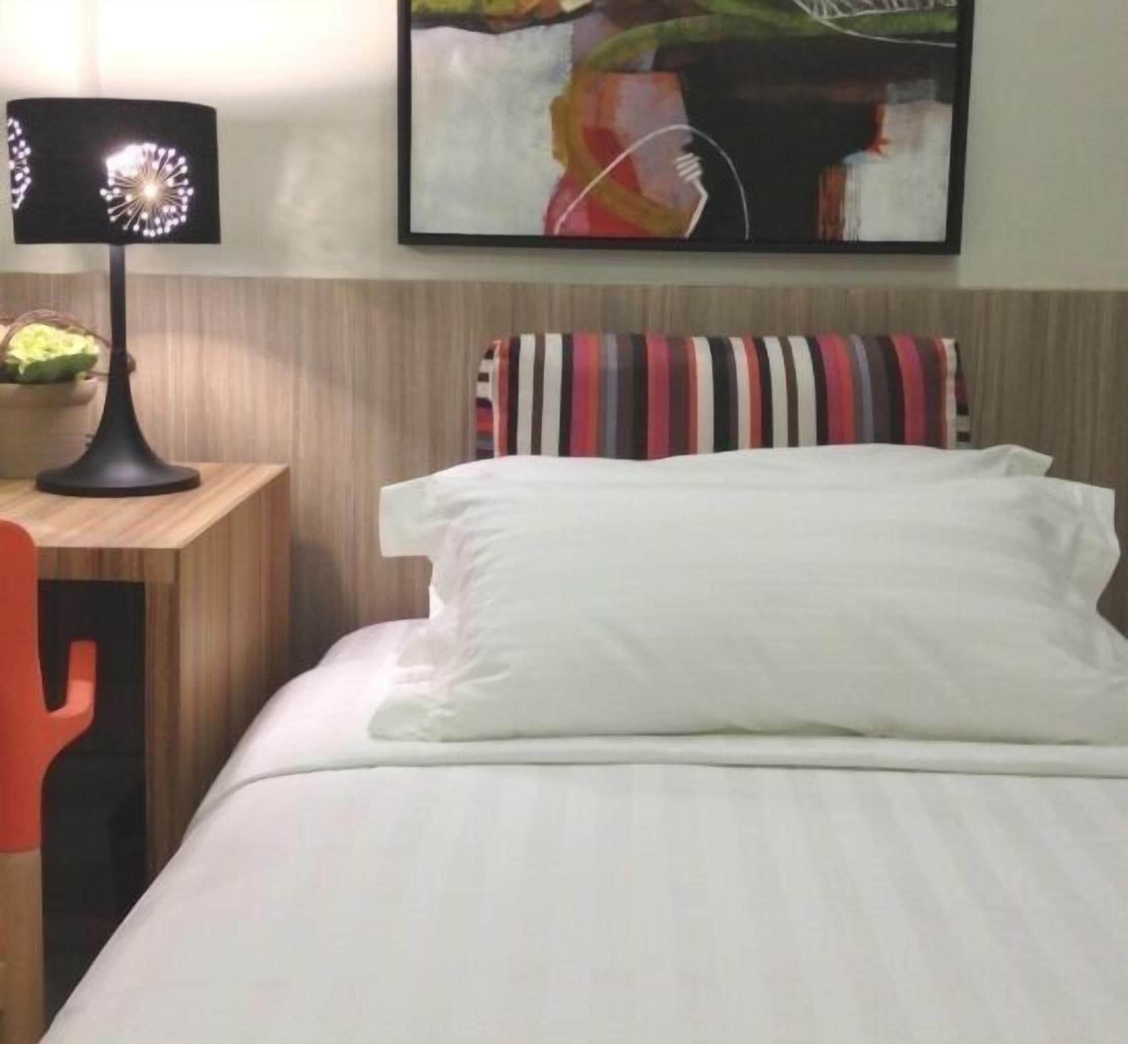 A neatly made bed with white linens, a black lamp on a wooden nightstand, and a colorful abstract painting above the bed.