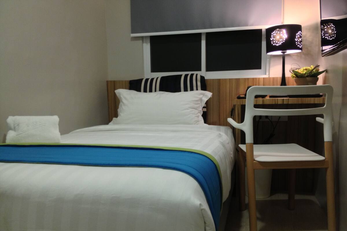 A neatly made bed with white bedding and a blue and green striped blanket. There is a white chair with a wooden backrest next to the bed. A black lamp with a floral design is on a table beside the bed.