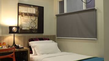 A hotel room with a bed, a desk with a lamp, a phone, and a window with a blind.