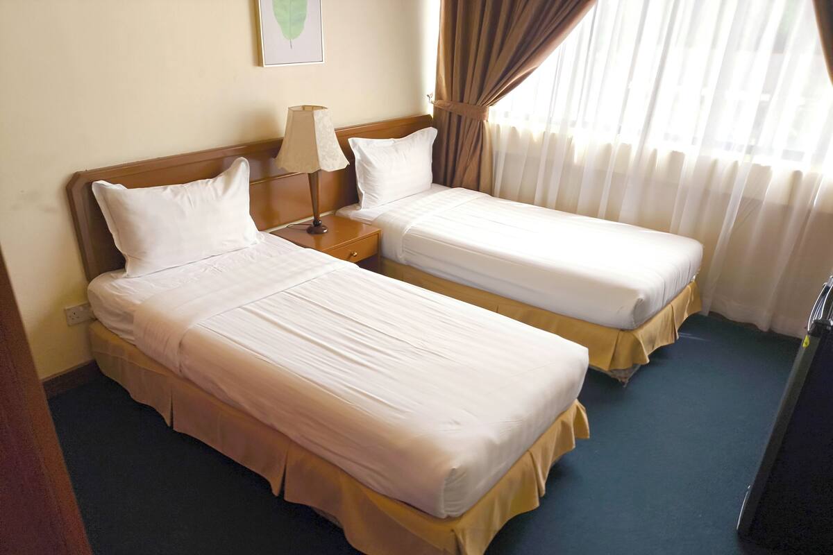 A hotel room with two single beds, a nightstand with a lamp, a framed picture on the wall, and a window with curtains.