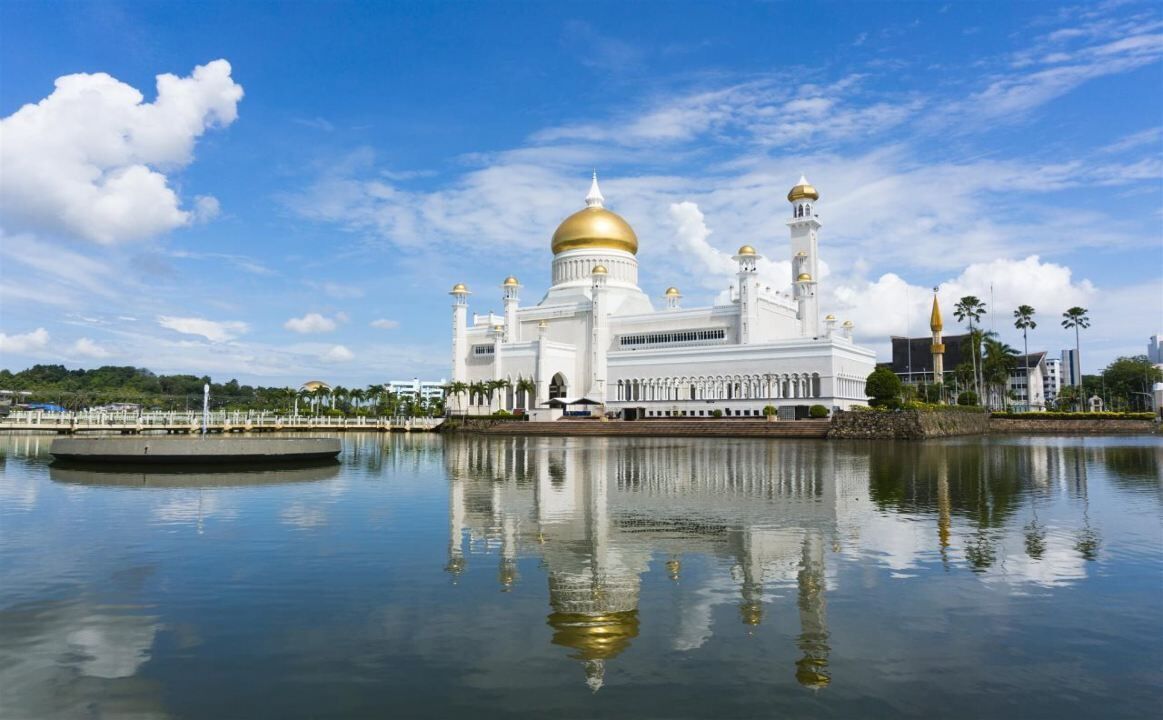 Un edificio grande y blanco con una cúpula dorada y múltiples minaretes, rodeado de agua y palmeras.