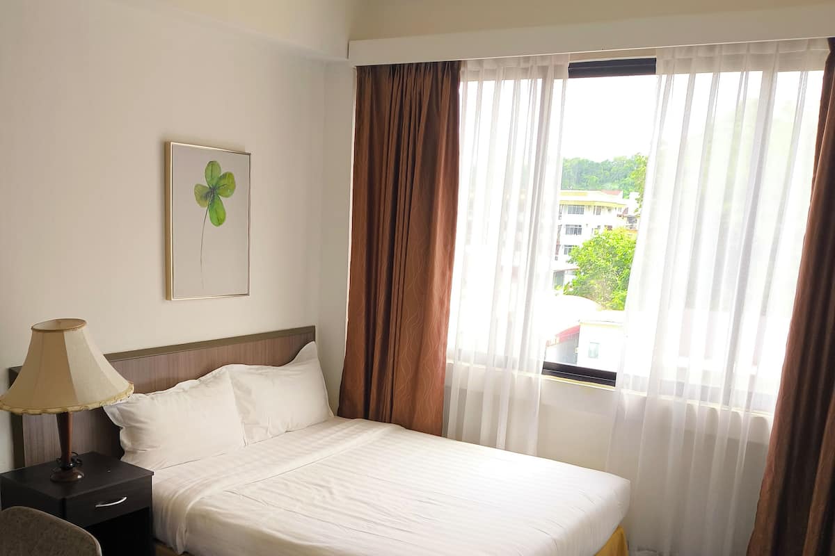 A hotel room with a large bed, a bedside table with a lamp, a chair, and a view of buildings through sheer curtains.