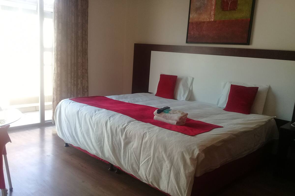 Habitación de hotel con una cama grande, almohadas rojas y blancas, una manta roja y un control remoto sobre la cama.