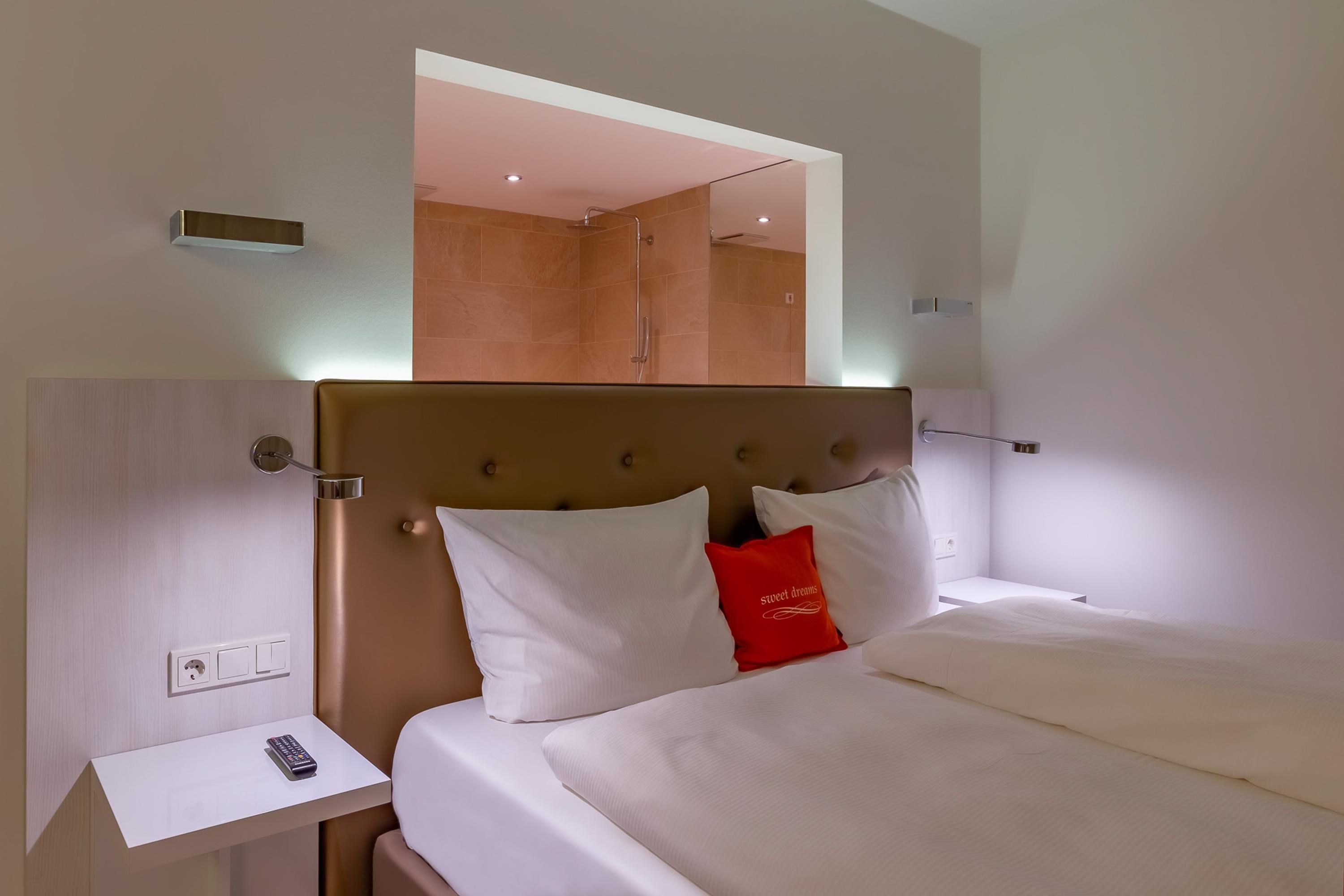 Habitación de hotel con cama, mesita de noche, espejo y zona de ducha.