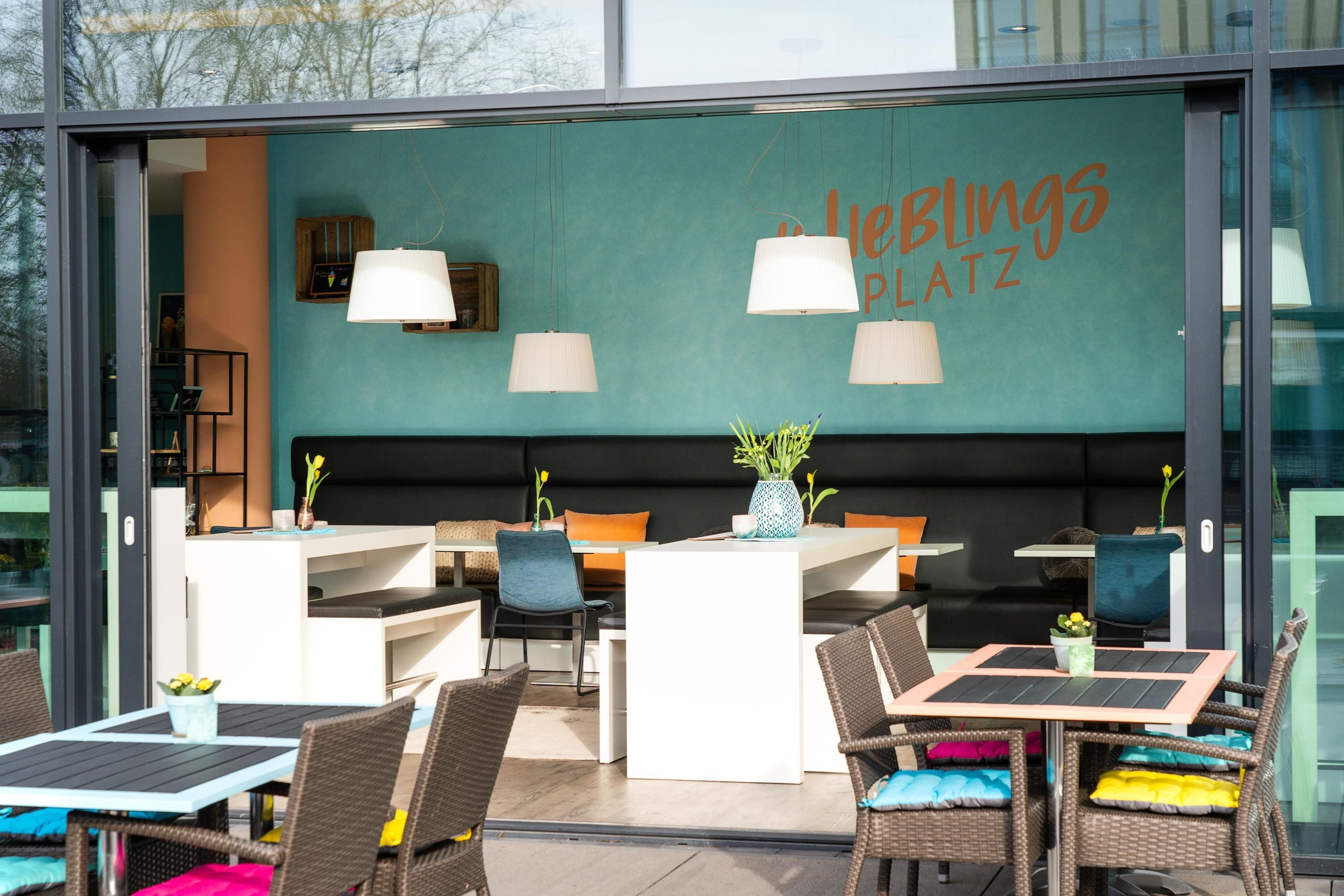 An outdoor seating area with tables and chairs, a teal wall with the text 'HOBBIES PLATZ', and pendant lights.