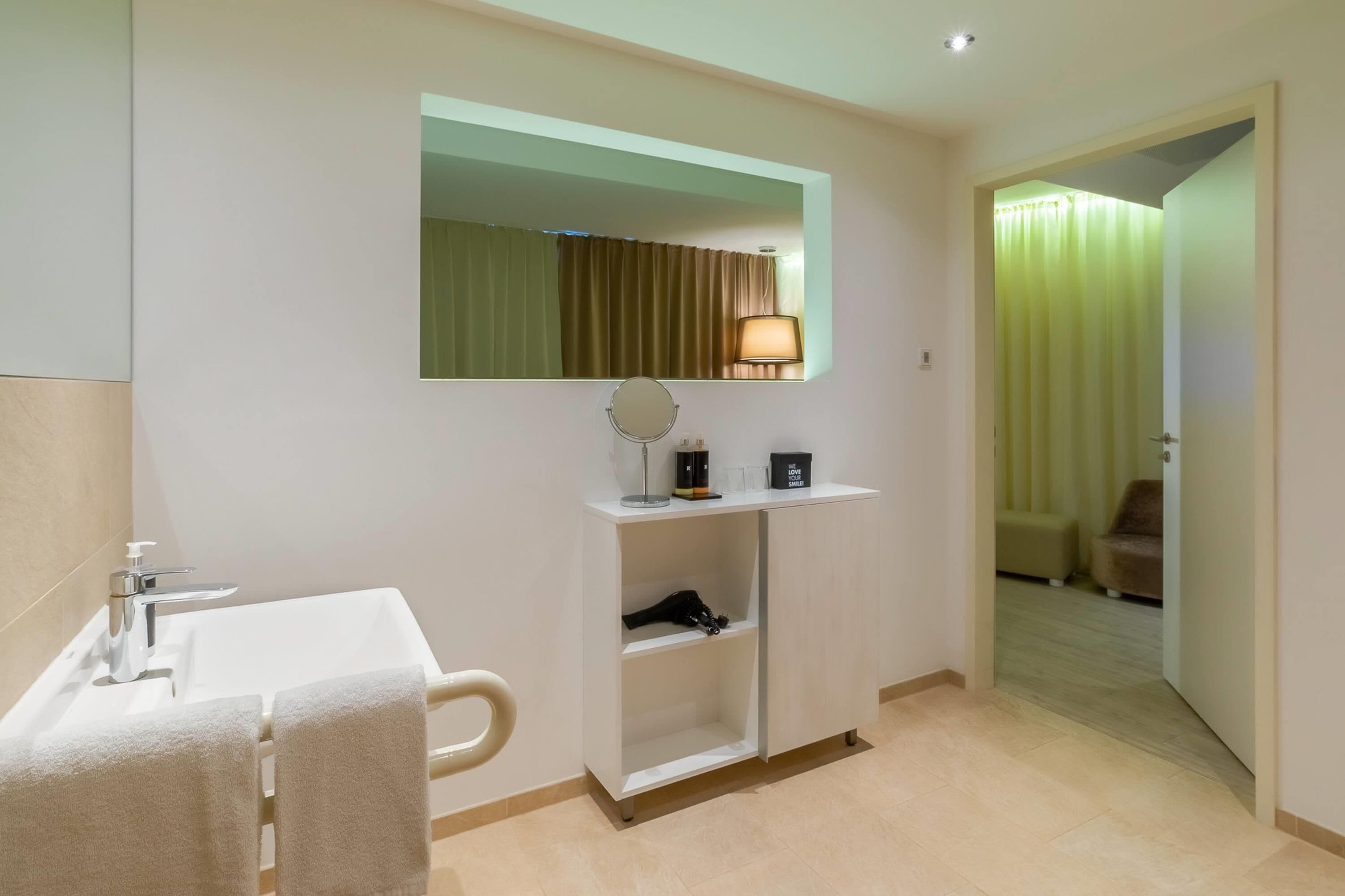 A hotel bathroom with a sink, mirror, towel rack, and a shelf with toiletries.