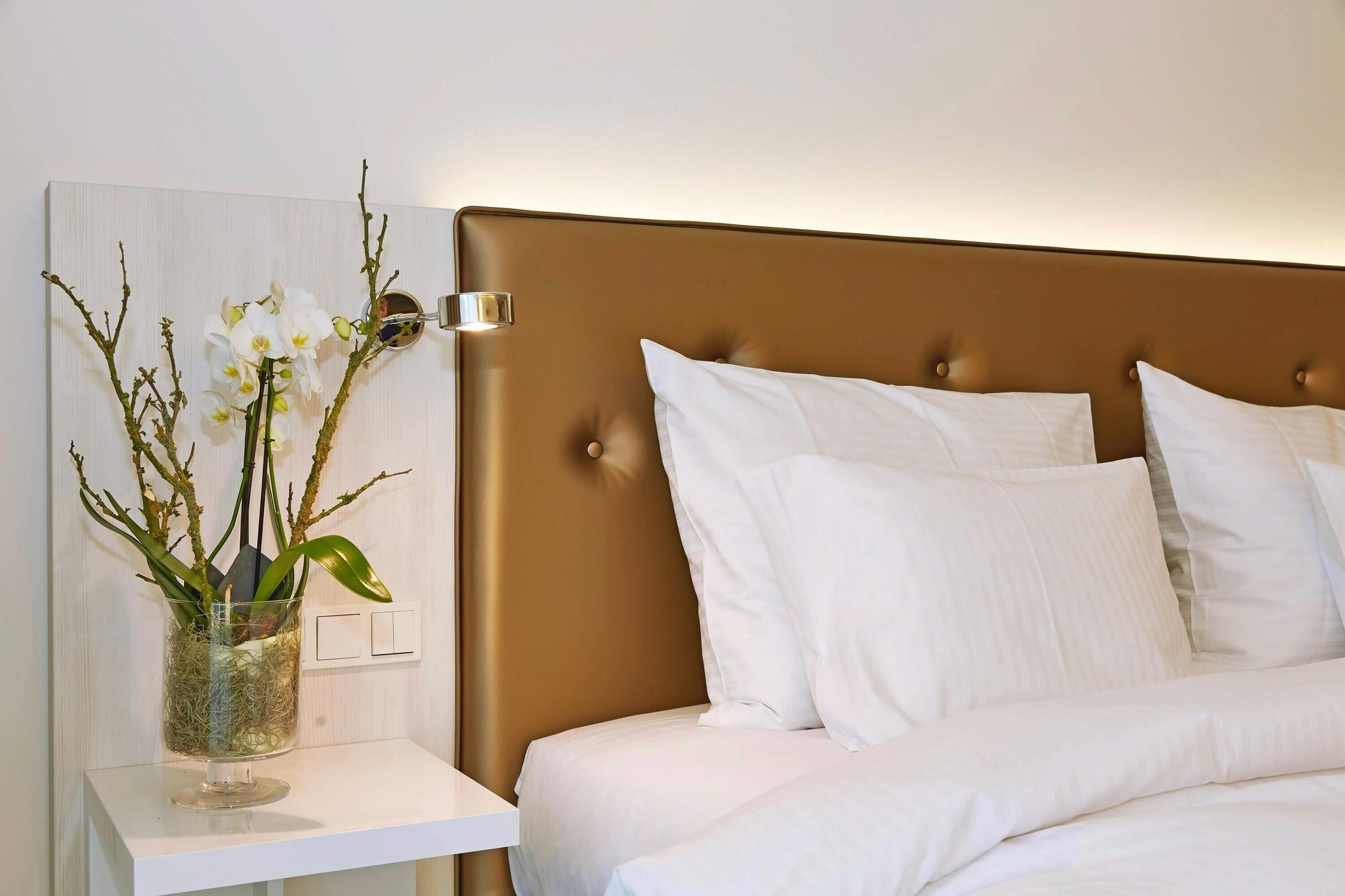 Un dormitorio con cama, ropa de cama blanca, una mesita de noche con un florero y una lámpara de pared.