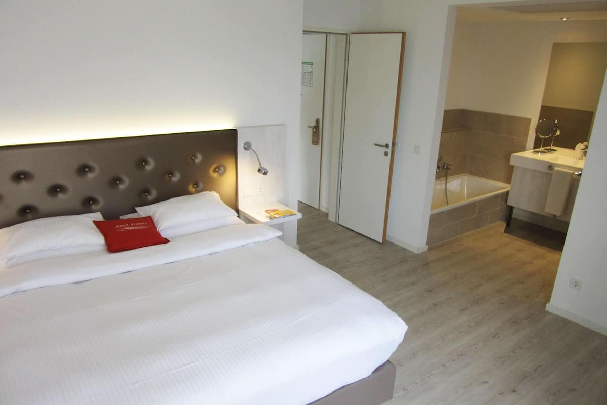 A hotel room with a bed, a nightstand, a red pillow, a bedside lamp, a door, and a bathroom with a bathtub and sink.