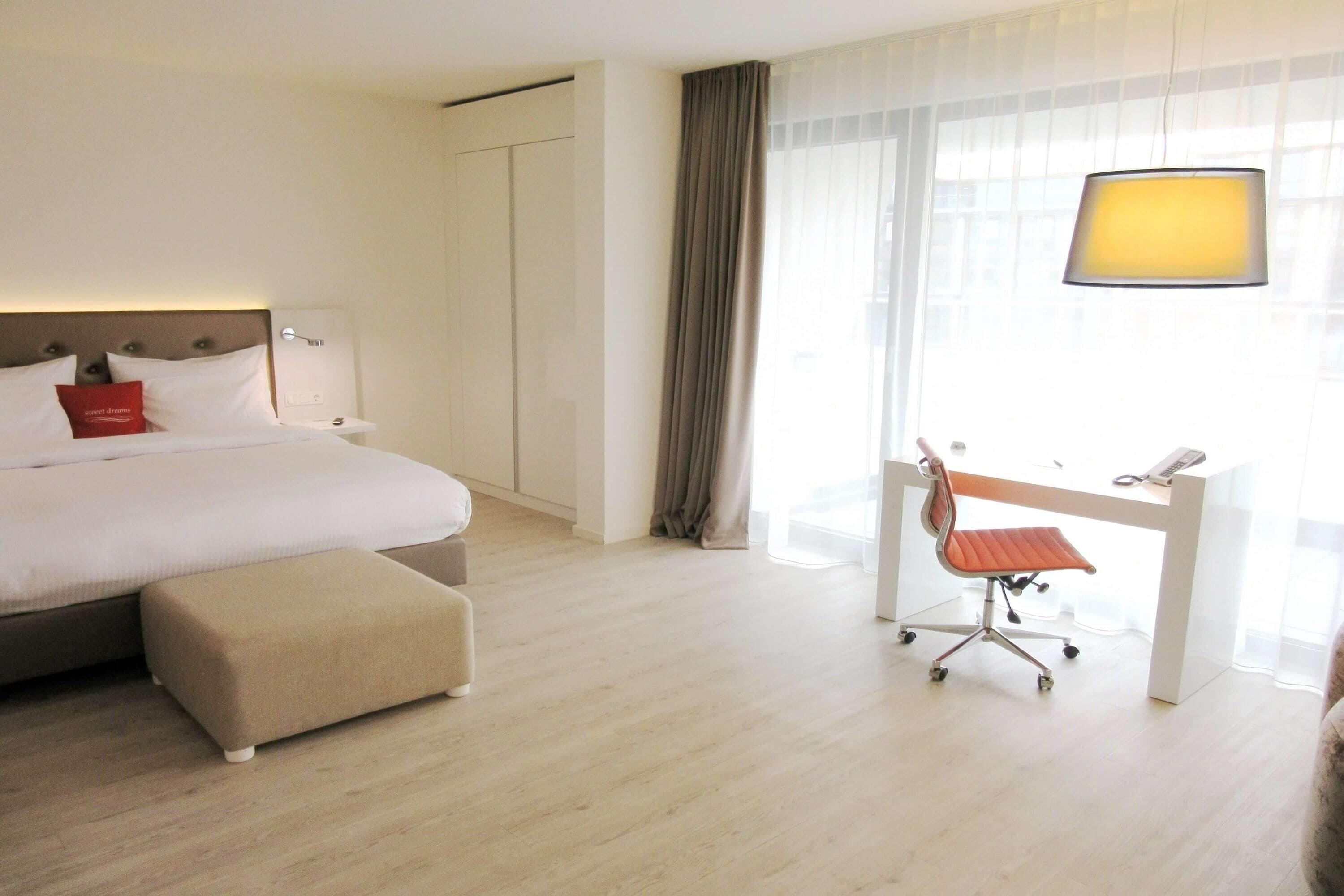 A hotel room with a bed, a desk with a chair, a lamp, and a window with curtains.