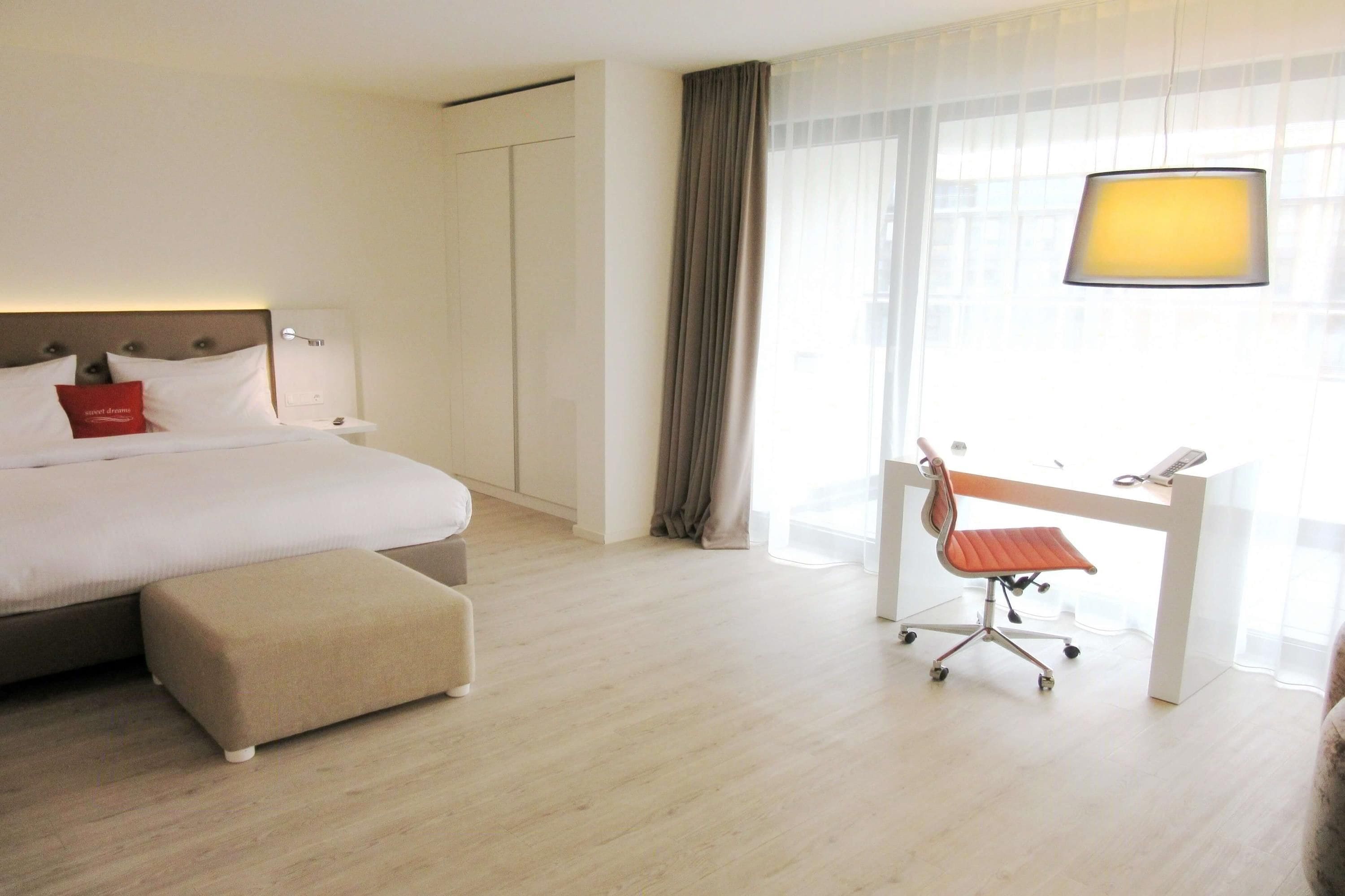 A hotel room with a bed, a desk with a chair, a lamp, and a window with curtains.