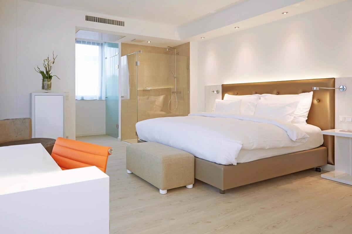 A modern bedroom with a large bed, a bedside table, a small desk, and a shower area.