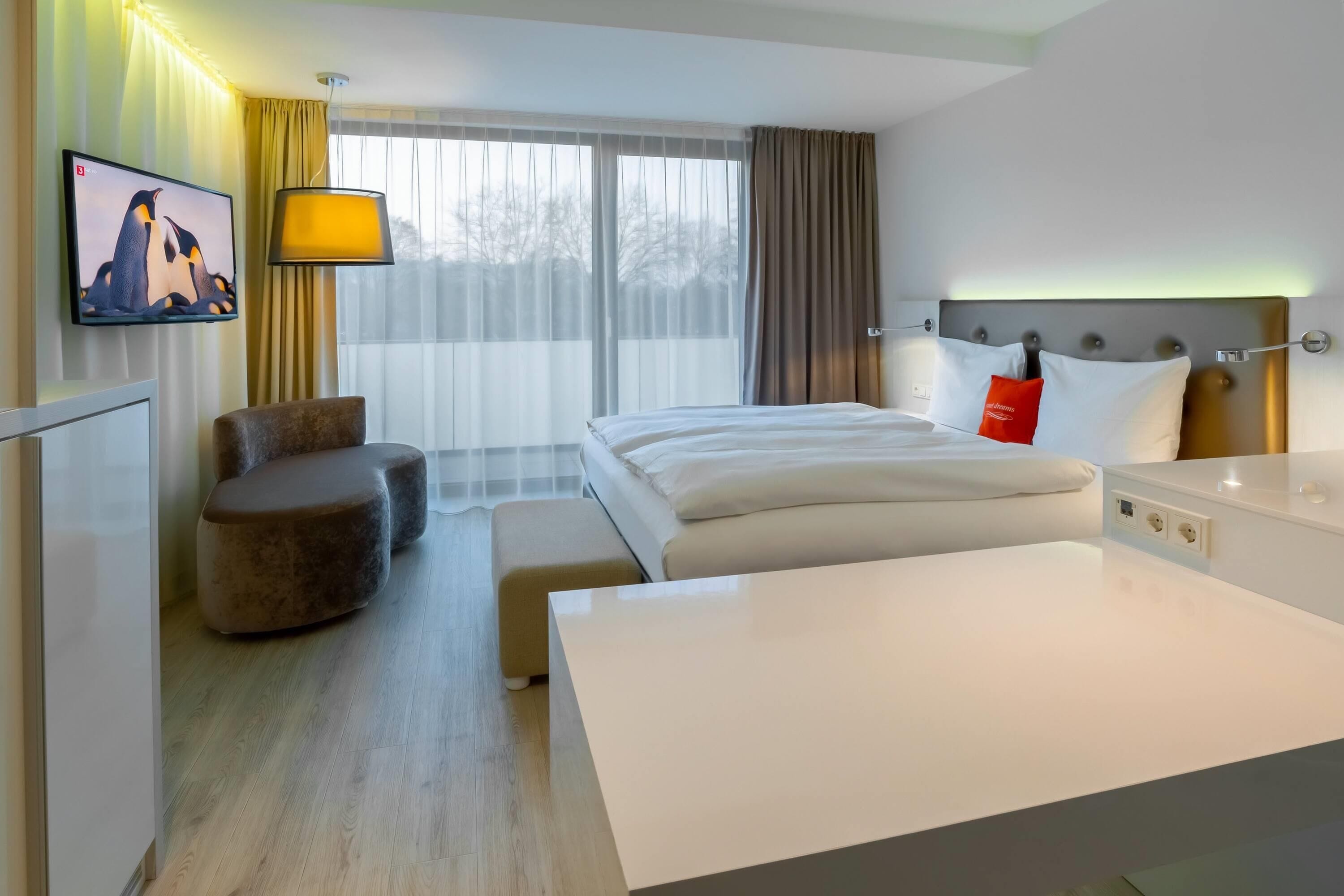 Quarto de hotel com cama, mesa de cabeceira, abajur e televisão na parede.