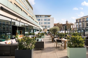 An outdoor seating area with tables and chairs, surrounded by greenery and overlooking a modern building with large windows.