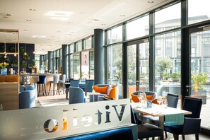A modern restaurant with large windows, blue and orange seating, and a visible bar area. - elaya hotel kleve, Trademark Collection by Wyndham (Kleve)