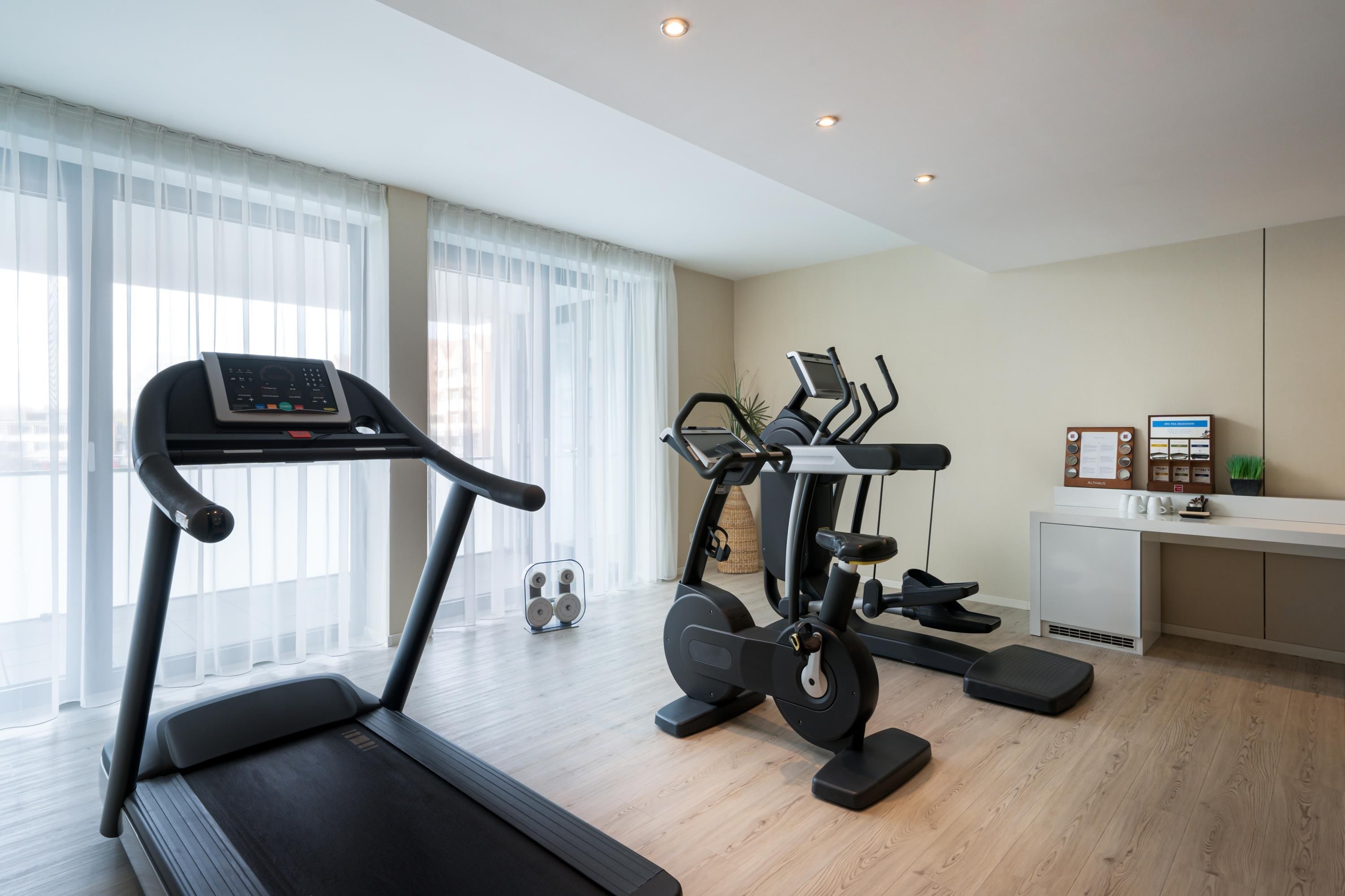A modern home gym with a treadmill, elliptical, and stationary bike, large windows with sheer curtains, and a clean, minimalist design.