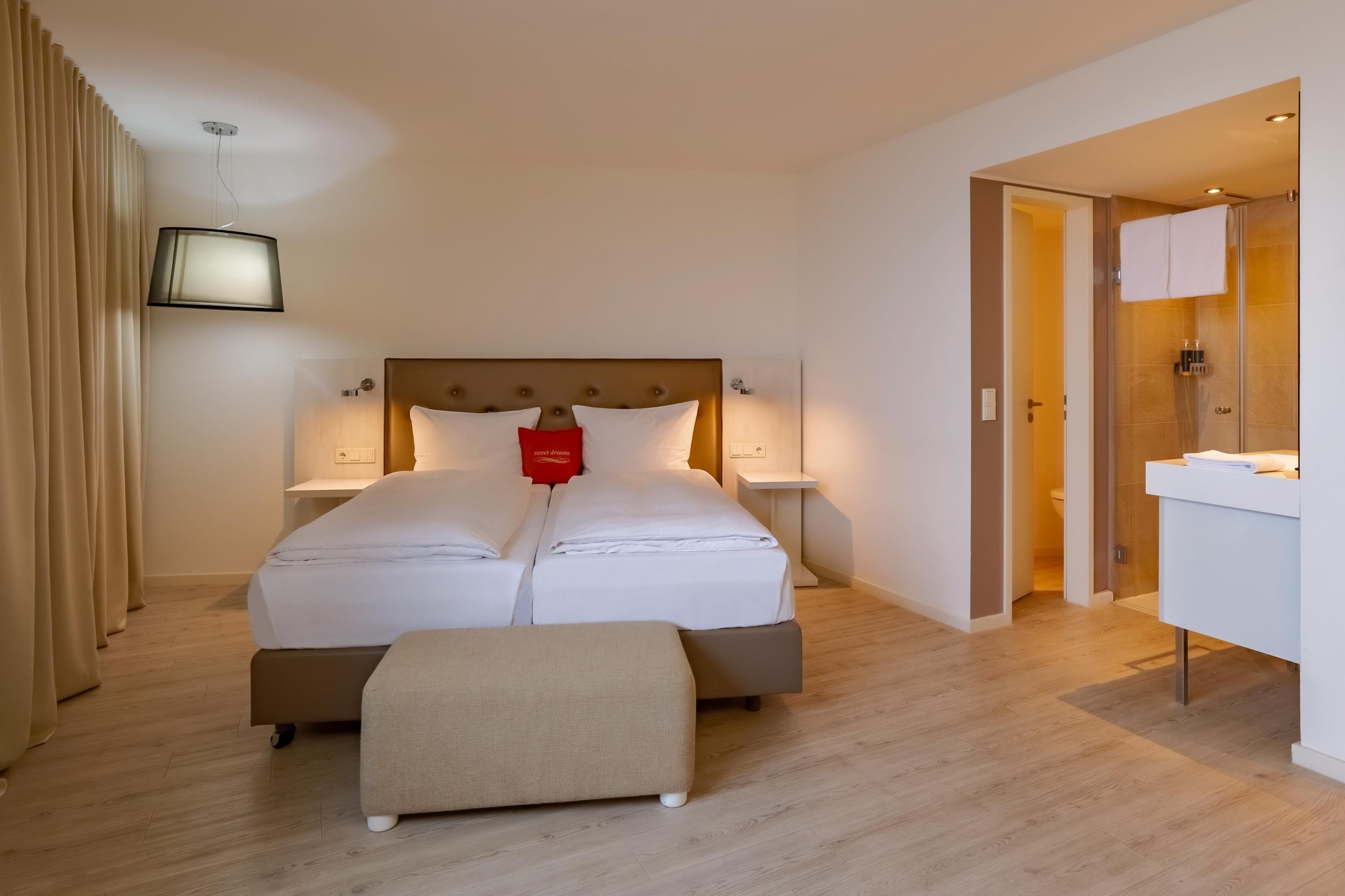 Una habitación de hotel con una cama grande, mesitas de noche, un banco y un baño visible a través de una puerta abierta.