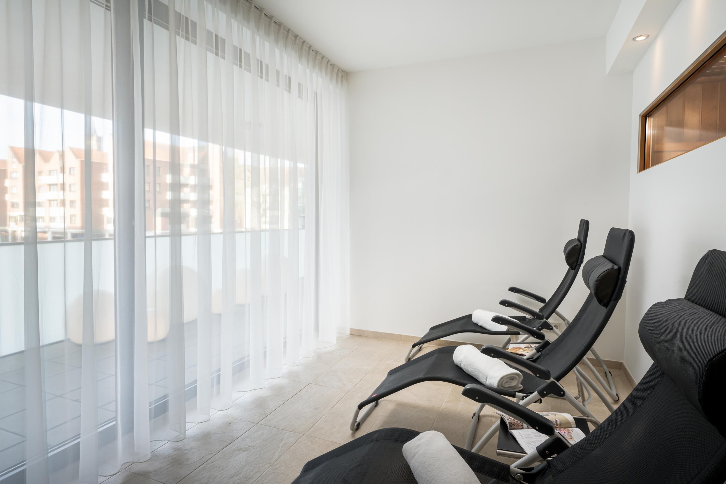A room with a balcony, large windows with sheer curtains, and several reclining chairs.