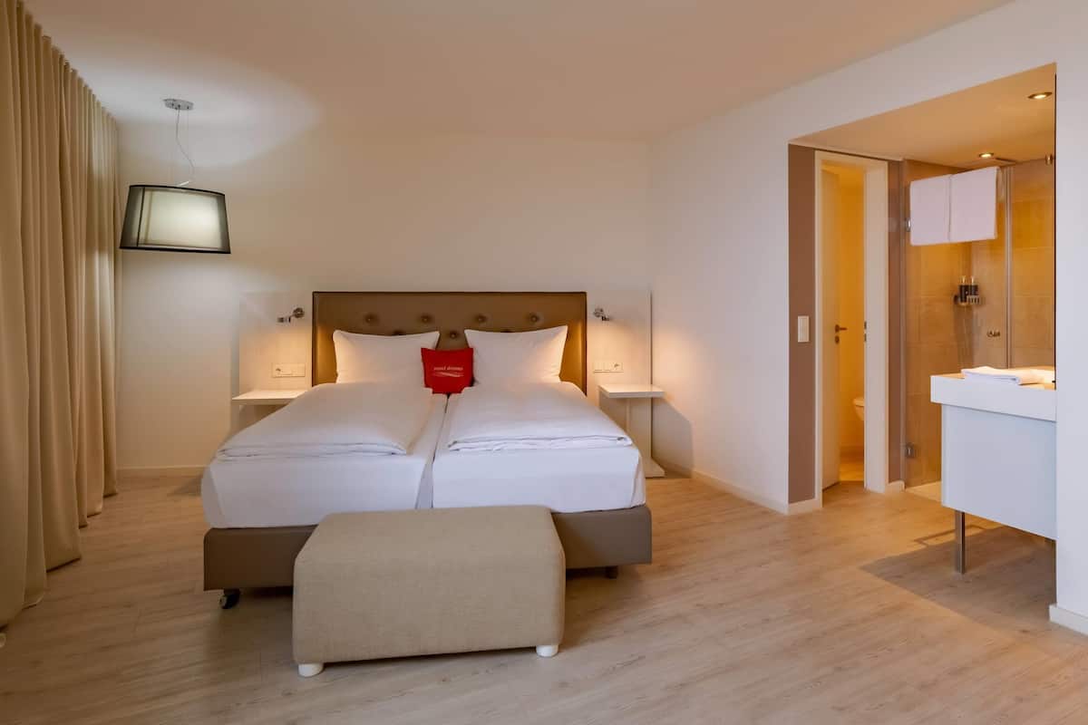 A hotel room with a large bed, bedside tables, a small table, a bench, a TV, and a bathroom visible through an open door.