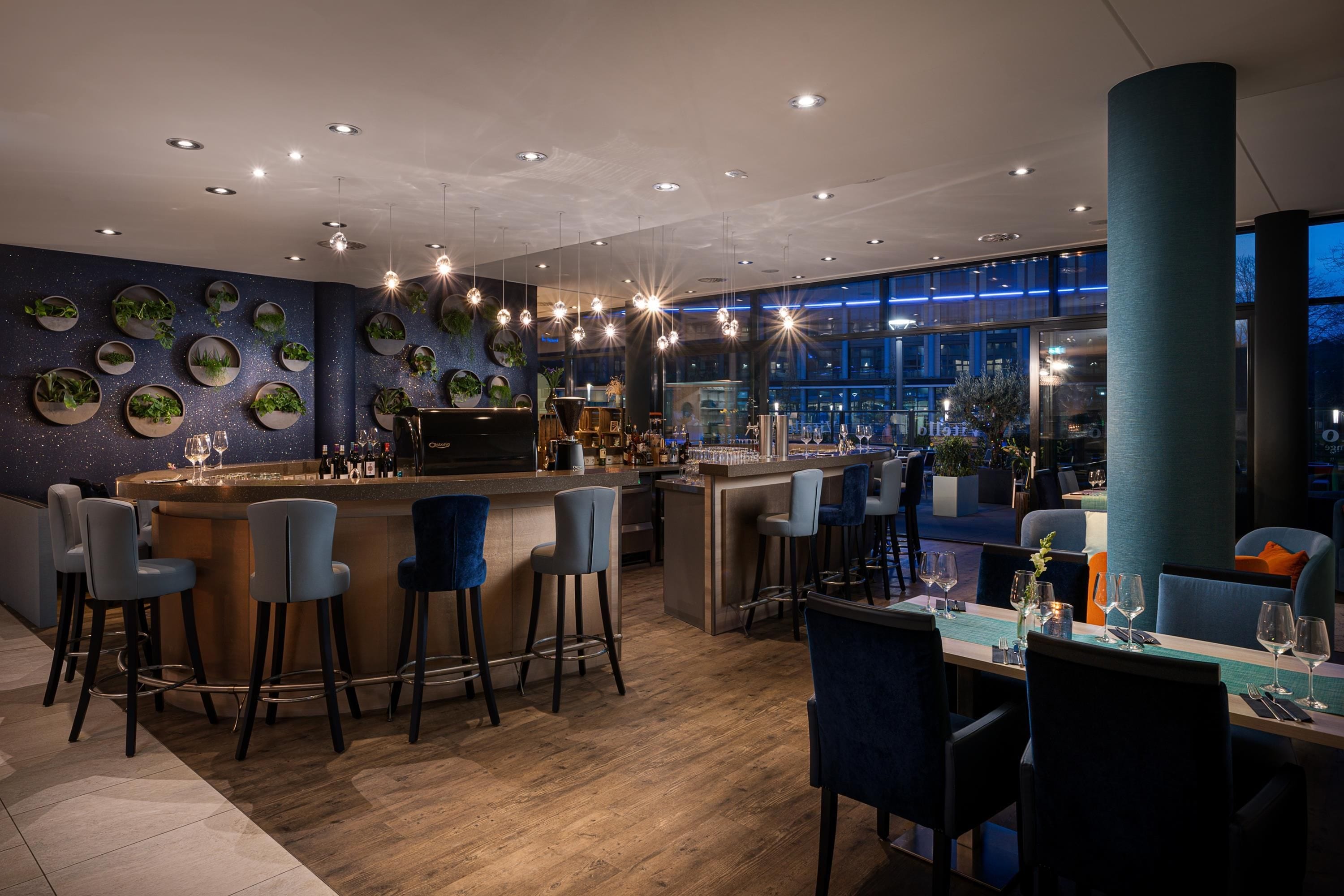 Un área de bar moderna con mostrador de madera, varios taburetes y una zona de estar con mesas y sillas.