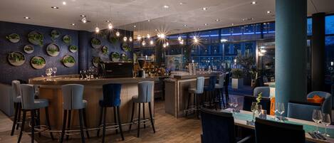 Un área de bar moderna con mostrador de madera, varios taburetes y una zona de estar con mesas y sillas.